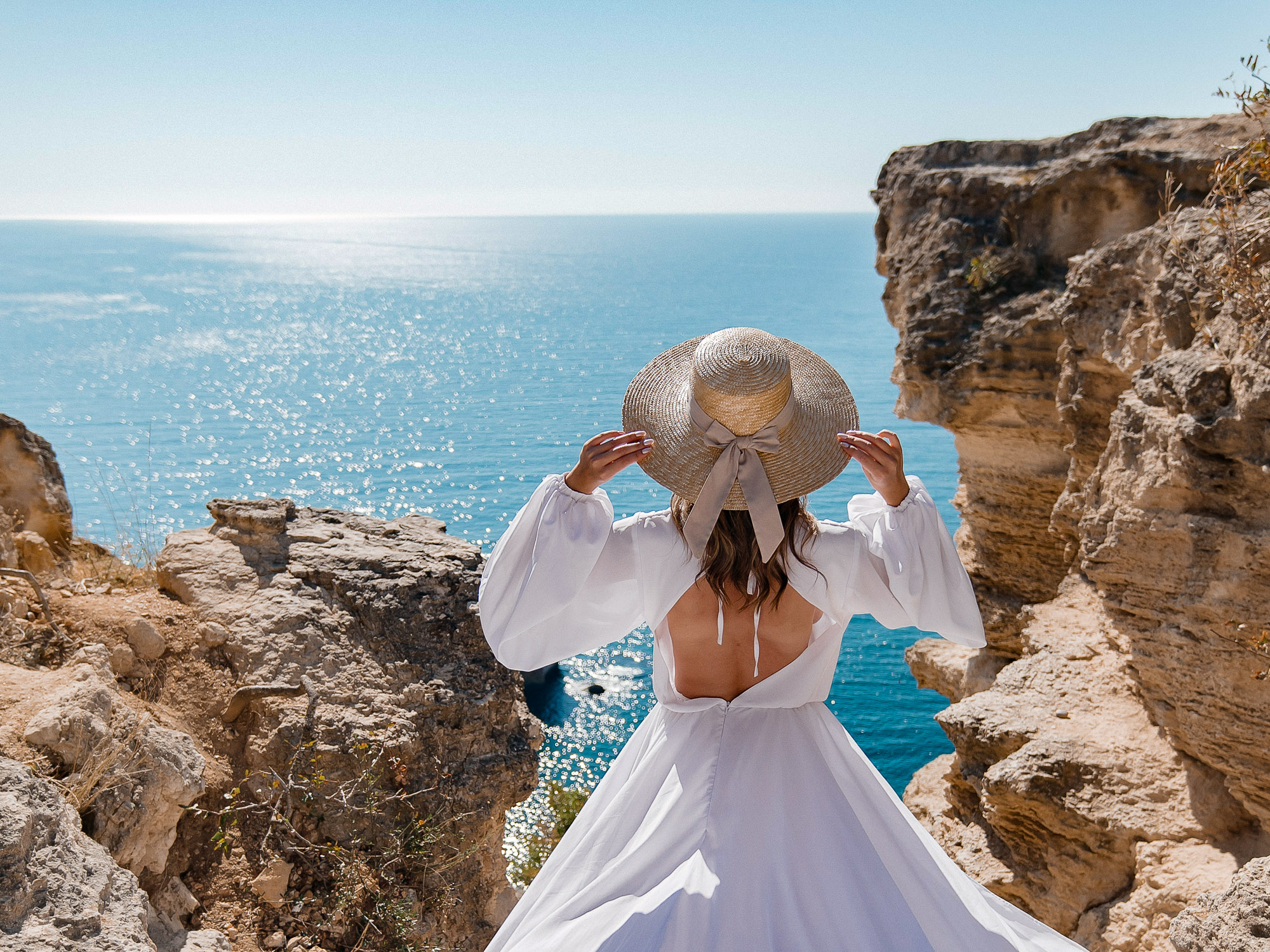 A woman in a white dress is walking on a rocky beach near the ocean. The scene is serene and peaceful, with the woman's dress flowing in the wind